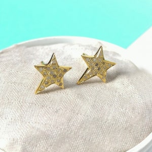 May include: A pair of gold star-shaped earrings with small white diamonds set in the metal. The earrings are displayed on a white velvet cushion inside a white box.