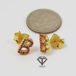 May include: A pair of gold-tone earrings with the letter "B" in pink cubic zirconia stones. The earrings are shown next to a US dime for size comparison.