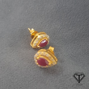 Könnte beinhalten: Ein Paar goldene Ohrringe mit einem tropfenförmigen roten Edelstein, der von kleineren weißen Edelsteinen umgeben ist.