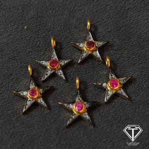 Puede incluir: Cinco amuletos de estrella plateados con una piedra preciosa roja en el centro de cada estrella. Las estrellas están bordeadas de oro y tienen pequeñas piedras preciosas blancas alrededor de los bordes.