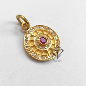 May include: A gold pendant with a round design featuring a red gemstone surrounded by smaller white gemstones. The pendant has a loop at the top for attaching to a chain.