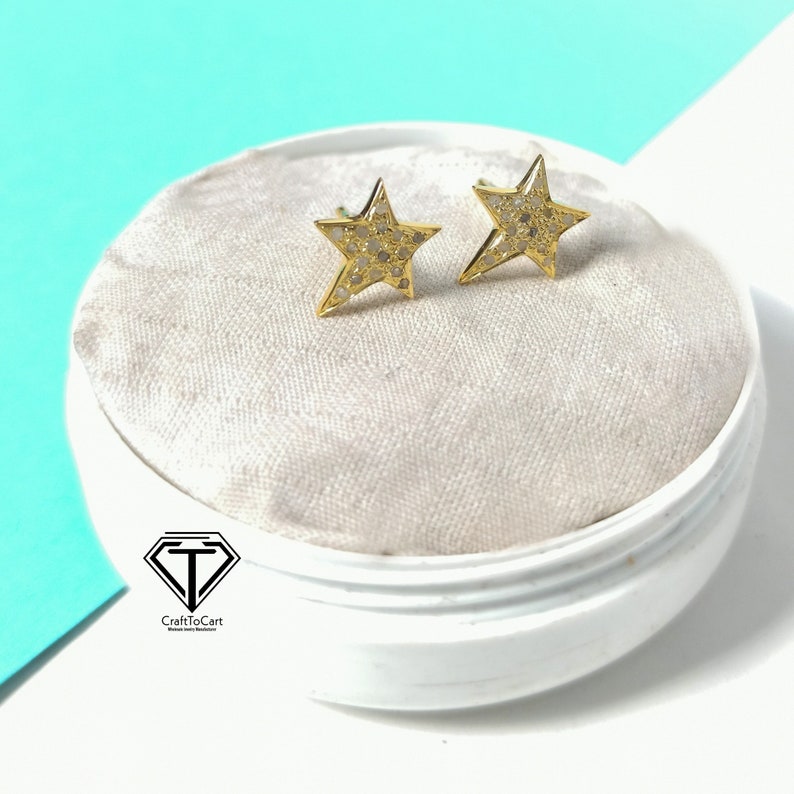 May include: A pair of gold star-shaped earrings with small white diamonds set in the metal. The earrings are displayed on a white velvet cushion inside a white box.