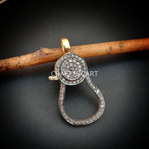 May include: A silver and gold clasp with a round, pave-set diamond design. The clasp is in the shape of a pear and has a small loop at the top.