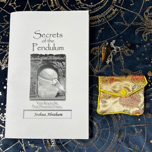May include: A white book titled "Secrets of the Pendulum" by Joshua Abraham. The book cover features a black and white illustration of an archway with a pendulum hanging from it. The book is on a blue background with a gold star pattern.