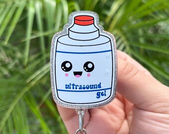 May include: A badge reel featuring a cartoon ultrasound gel bottle design. The bottle is white with a red cap and blue accents, and the words "ultrasound gel" are printed on the front. The badge reel has a silver glitter border and a retractable cord.