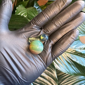 May include: A collection of glass marbles in various shapes and colors. The marbles include a clear, rectangular piece, a clear marble with a yellow tint, and a green and brown marble. The marbles are held in a black gloved hand.