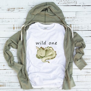 May include: White t-shirt with a green cartoon dragon and the text "wild one" printed on it. The t-shirt is laying on a wooden surface with a green hooded sweatshirt.
