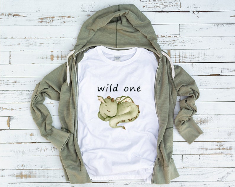 May include: White t-shirt with a green cartoon dragon and the text "wild one" printed on it. The t-shirt is laying on a wooden surface with a green hooded sweatshirt.