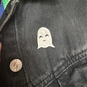 May include: A small, white ghost-shaped pin with a smiling face is attached to a black denim jacket. The pin has a simple design with blue eyes and a black mouth. A silver button with the text "LOW TOPS" is also visible.