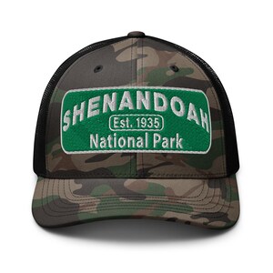 May include: A camouflage print trucker hat with a black mesh back and a rectangular green embroidered patch on the front. The patch features white text that reads SHENANDOAH, Est. 1935, and National Park, centered on the front panels of the cap.