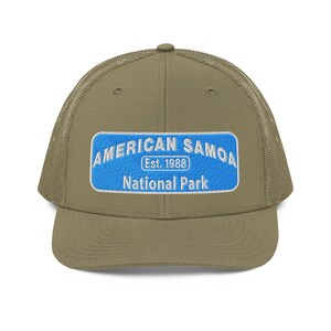 May include: A khaki-colored trucker hat with a blue rectangular patch featuring the text "AMERICAN SAMOA Est. 1988 National Park" in white lettering. The hat has a mesh back and a curved brim.