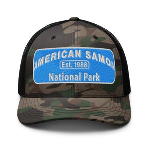 May include: A camouflage trucker hat with a black mesh back and a curved brim. The front panel features a blue patch embroidered with "AMERICAN SAMOA", "Est. 1988", and "National Park" in white lettering. The hat is a classic accessory.