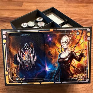 May include: A rectangular black game box lid featuring a metallic silver emblem on the left and a female cyborg figure against a fiery space background on the right. The open box base behind it contains several circular game tokens and cards.
