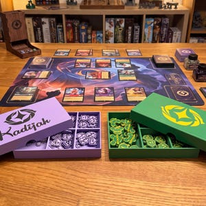May include: A tabletop game setup featuring a playmat with cards, a wooden dice tower, and two rectangular storage boxes. One lavender box is labeled Kadijah and contains purple tokens, while a green box holds green tokens with yellow symbols.
