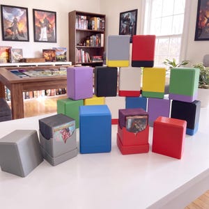 May include: A collection of rectangular plastic trading card deck boxes in various solid colours including red, blue, green, purple, black, grey, and white. Some boxes are closed, while others are open, revealing stacks of collectible game cards.