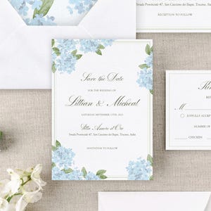 Puede incluir: Juego de invitaciones de boda blancas con detalles florales de hortensias azules. La invitación dice "Save the Date" para Lillian & Micheal, sábado 13 de septiembre de 2025. Incluye un sobre y una tarjeta de respuesta.