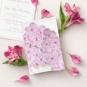 Könnte beinhalten: Ein helllila Hochzeitskarten-Set mit floralen Ausschnitten und einer passenden Karte. Die Einladung enthält die Namen Gabriella Ellen Rosen und Oliver Peter Henderson. Rosa Blütenblätter und Blüten sind um die Einladung verstreut.