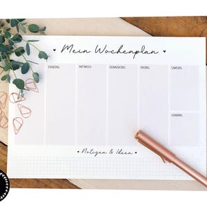 Könnte beinhalten: Ein weißer Wochenplaner mit dem Text "Mein Wochenplan" und "Notizen & Ideen" in schwarzer Schrift. Der Planer hat Abschnitte für jeden Wochentag. Ein roségoldener Stift liegt auf dem Planer, mit roségoldenen Büroklammern und Grünzeug im Hintergrund.
