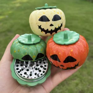 Puede incluir: Tres molinillos de hierbas con forma de calabaza y caras de jack-o'-lantern. Uno es crema, otro verde y otro naranja, todos con pintura moteada. El molinillo verde está abierto, revelando un interior metálico con dientes afilados.