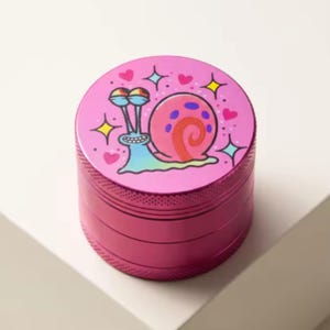 Puede incluir: Un molinillo de hierbas cilíndrico de metal rosa con un gráfico de un caracol de dibujos animados con concha rosa y cuerpo azul. La tapa superior está decorada con pequeños corazones rosas y estrellas de cuatro puntas amarillas y azules.
