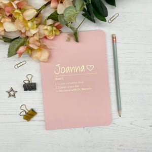 May include: A light pink notebook featuring the name Joanna followed by the word noun and a list of three personal traits printed in white text. A grey pencil and several metal paper clips are arranged around the notebook.