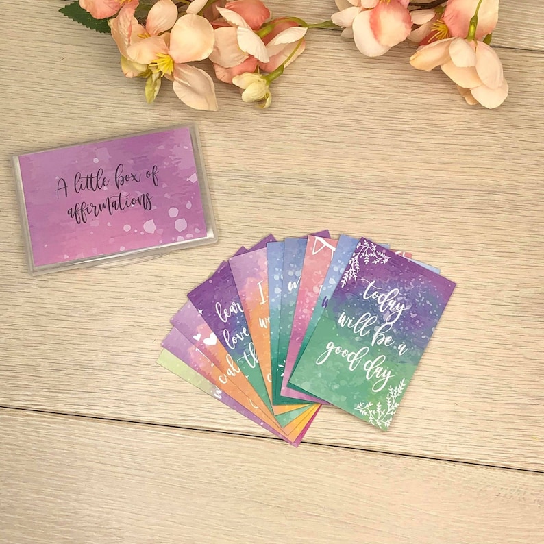 K&ouml;nnte beinhalten: Ein Set von Affirmationskarten mit Aquarellhintergr&uuml;nden in Lila, Blau und Gr&uuml;n. Die Karten haben inspirierende S&auml;tze in wei&szlig;er Schrift. Das Set enth&auml;lt eine transparente Plastikbox mit dem Text "A little box of affirmations".