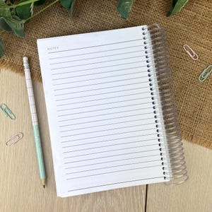 May include: A white spiral-bound notebook with lined pages and the word NOTES printed at the top. A pencil with a white and light green grid pattern and a silver ferrule sits beside the notebook. Four small metal paper clips in pink and light blue are scattered around the notebook.