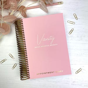 May include: A pink appointment diary with a gold spiral binding. The cover features the word "Vanity" in elegant script, along with "NAILS BROWS & BEAUTY" and "APPOINTMENT DIARY" in gold lettering. Gold paper clips and a pencil are scattered around the diary.