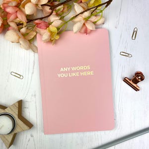 May include: A pale pink notebook with the gold text "ANY WORDS YOU LIKE HERE". The notebook is on a white wooden surface, with paperclips, a decorative clip, and artificial flowers arranged around it.