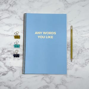 May include: A pale blue spiral-bound notebook with gold text reading "ANY WORDS YOU LIKE". A yellow pencil and three binder clips in gold, teal, and black are also present.