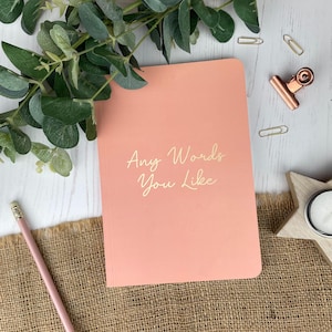 May include: A coral-coloured notebook with the text "Any Words You Like" in a cursive font. The notebook is surrounded by stationery items, including a pencil, paper clips, and a decorative clip. Green foliage adds a natural touch.