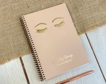 May include: A spiral-bound appointment book with a light pink cover featuring gold foil graphics of closed eyes with long eyelashes. The text "Le Luz Beauty" and "APPOINTMENTS" is printed in gold foil. A metallic rose gold pen lies beside the book.