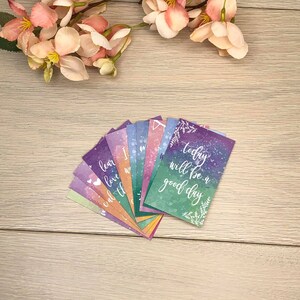 K&ouml;nnte beinhalten: Eine Sammlung farbenfroher Karten mit inspirierenden Zitaten, die auf einer hellen Holzoberfl&auml;che aufgef&auml;chert sind. Die Karten haben Aquarellhintergr&uuml;nde in Lila, Blau und Gr&uuml;n mit wei&szlig;er Schrift. Rosa und pfirsichfarbene Blumen sind im Hintergrund.