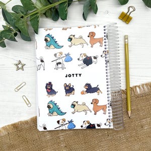 May include: A spiral-bound notebook with a white cover featuring a repeating pattern of various dog breeds in different outfits and poses. The word JOTTY is printed in black in the center. The notebook is accompanied by a yellow wooden pencil.