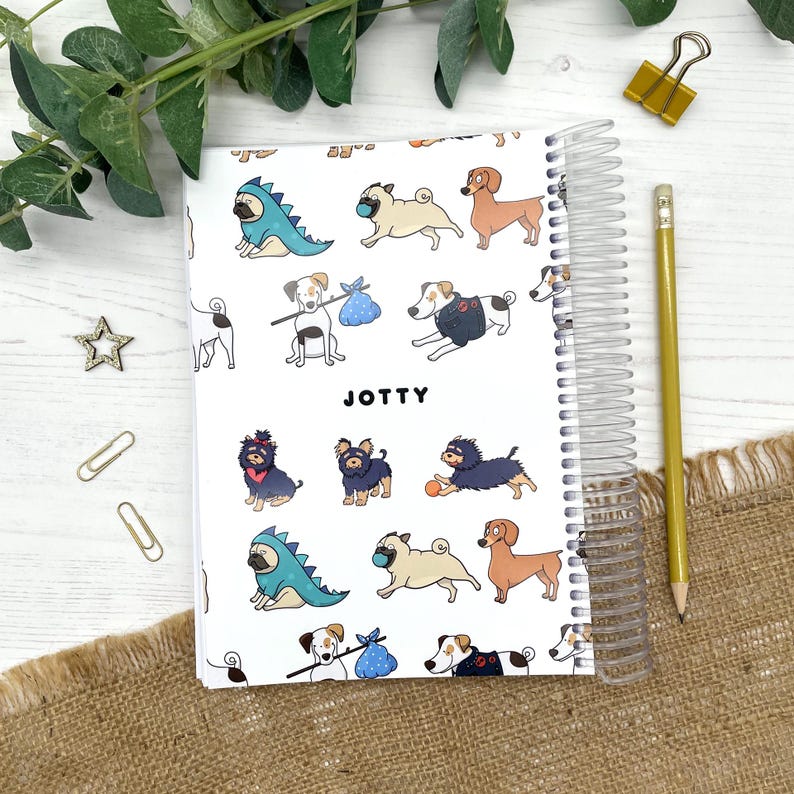 May include: A spiral-bound notebook with a white cover featuring a repeating pattern of various dog breeds in different outfits and poses. The word JOTTY is printed in black in the center. The notebook is accompanied by a yellow wooden pencil.