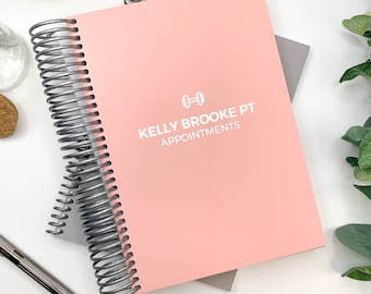 May include: A pink spiral-bound planner with the text "KELLY BROOKE PT APPOINTMENTS" and a dumbbell icon in white. The planner is stacked on top of a gray planner. A pen, a small decorative vase, and greenery are also visible.