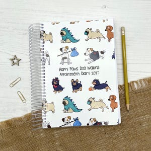 May include: A spiral-bound appointment diary with a white cover featuring a repeating pattern of various cartoon dogs, including pugs in dinosaur costumes. The text reads Happy Paws Dog Walking Appointment Diary 2027. A yellow pencil is placed beside it.