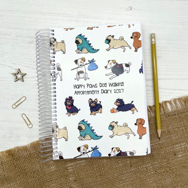May include: A spiral-bound appointment diary with a white cover featuring a repeating pattern of various cartoon dogs, including pugs in dinosaur costumes. The text reads Happy Paws Dog Walking Appointment Diary 2027. A yellow pencil is placed beside it.