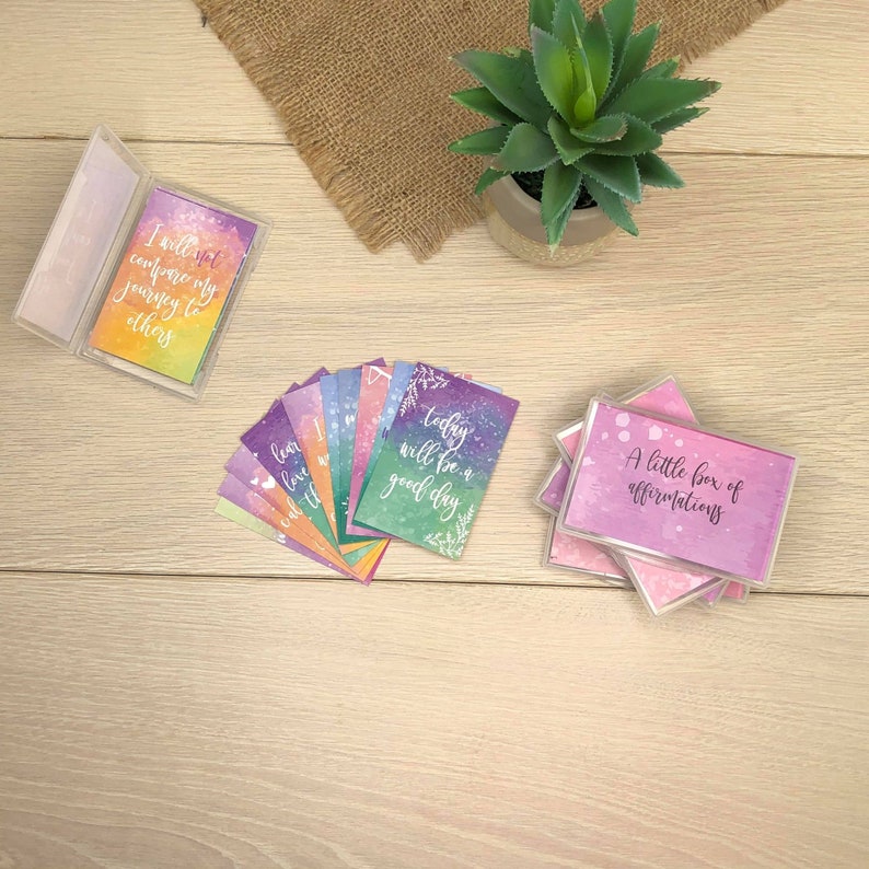 K&ouml;nnte beinhalten: Eine Sammlung von Affirmationskarten mit Aquarellhintergr&uuml;nden und inspirierenden Zitaten. Die Karten befinden sich in transparenten Kunststoffh&uuml;llen und sind auf einer hellen Holzoberfl&auml;che ausgebreitet. Eine Karte lautet: "Ich werde meine Reise nicht mit anderen vergleichen."