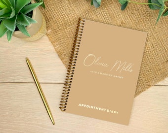 May include: A tan spiral-bound appointment diary with gold wire binding. The cover features the white text Olivia Mills, HAIR & MAKE UP ARTIST, and APPOINTMENT DIARY. A gold pen is positioned to the left of the diary on a light wood surface.