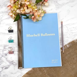 May include: A light blue spiral-bound diary with the text Bluebell Balloons and 2027 DIARY printed on the cover. The diary features a dark bronze coil binding and is accompanied by three small metal binder clips and a single wooden pencil.