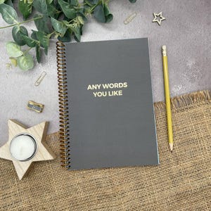 May include: A grey spiral-bound notebook with gold lettering reading "ANY WORDS YOU LIKE". A yellow pencil lies next to the notebook, and a wooden star-shaped candle holder is in the lower left corner. The background includes greenery and stationery.