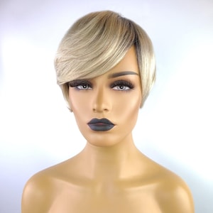 May include: A short, layered wig with a side-swept bang. The wig features a blend of blonde and light brown tones, creating a natural look. The wig is styled on a mannequin head with dark lipstick and long eyelashes. The background is white.