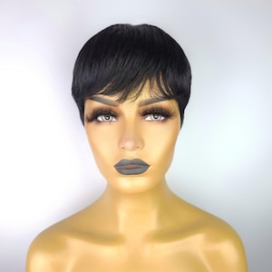 May include: A black pixie cut wig with a short, layered style and side-swept bangs. The wig is styled on a mannequin head with full makeup, including dark lipstick and long eyelashes. The background is a plain white.