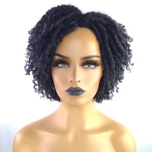 May include: A black, short, curly wig styled on a mannequin head. The wig features a natural-looking hairline and a textured, voluminous style. The mannequin has dramatic eye makeup and dark lipstick, set against a white background.