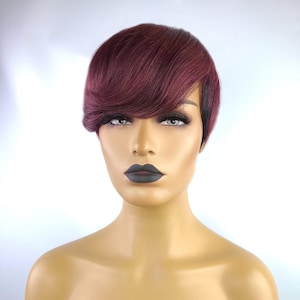 May include: A burgundy-colored pixie cut wig with a side-swept bang. The wig is styled on a mannequin head with dark lipstick and long eyelashes. The wig is a short, layered style.
