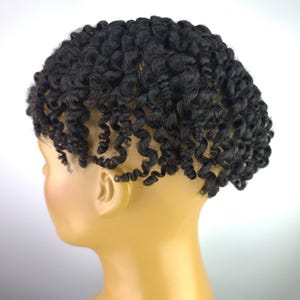 Op de afbeelding: Een zwarte, krullende pruik gestyled op een mannequinhoofd. De pruik heeft strakke, spiraalvormige krullen en is ontworpen om het hoofd te bedekken. Het mannequinhoofd heeft een lichte huidskleur, de achtergrond is wit.