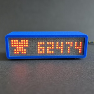 May include: A blue rectangular digital clock showing the numbers "X 62474" in orange LED lights. The clock has a blue frame and a grey screen. A black and yellow striped cord extends from the back.