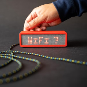 Puede incluir: Un dispositivo rectangular rojo muestra la palabra "WiFi?" en letras pixeladas rojas. Una mano se extiende hacia el dispositivo. Un cable estampado con detalles en azul y amarillo está en primer plano, sobre un fondo oscuro.