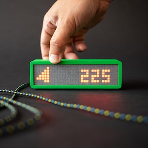 Könnte beinhalten: Ein grünes rechteckiges Digitaldisplay mit der Zahl "225" und einem Signal-Symbol. Das Display wird von einer Hand vor einem dunklen Hintergrund gehalten. Eine gemusterte Schnur befindet sich im Vordergrund.